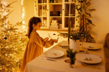 Noel süslemeli yemek masası, bulaşıklar, bardaklar ve mumlar, servis. Genç kız mum yakıyor.