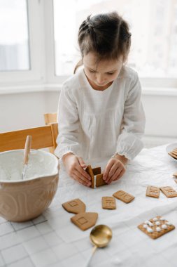 Beyaz bluz giyen sevimli kahverengi saçlı kız mutfakta zencefilli kurabiye evleriyle süslenmiş. Noel tatlısı, popüler Noel süslemeleri. Zencefilli kurabiye evi - klasik bayram geleneği