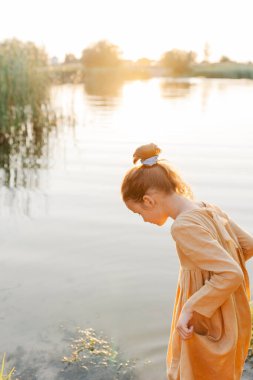 Curly caucasian girl wearing natural linen dress standing turned back in lake water, summertime. Coutryside living. Summer vibes. The feeling of summer.