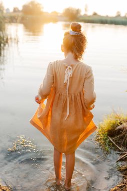 Curly caucasian girl wearing natural linen dress standing turned back in lake water, summertime. Coutryside living. Summer vibes. The feeling of summer.