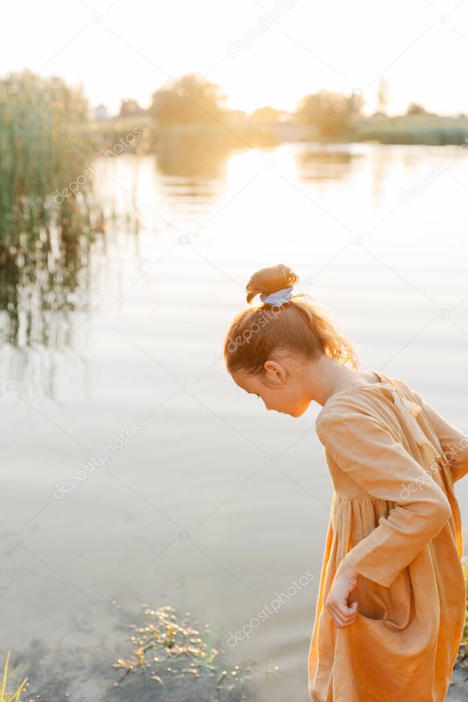 Curly caucasian girl wearing natural linen dress standing turned back in lake water, summertime. Coutryside living. Summer vibes. The feeling of summer.