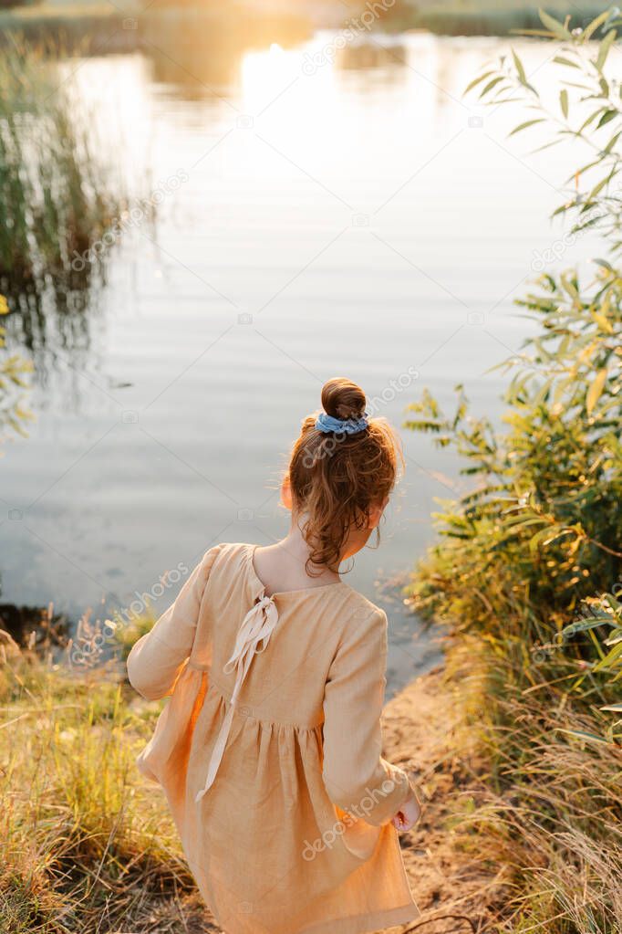 Curly caucasian girl wearing natural linen dress standing turned back for the lake, summertime. Coutryside living. Summer vibes. The feeling of summer.