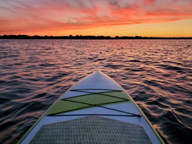 Gün batımının renkli ışığıyla suda yüzen kürek tahtası alacakaranlıkta bir gölün dalgalarından yansıyor.