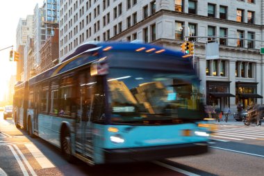 New York şehri otobüsü 23. Cadde 'den Manhattan' a doğru gidiyor. Arka plandaki binaların arasında güneş ışığı parlıyor.
