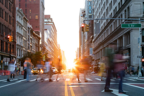 Busy street scene is crowded with people at an intersection on Fifth Avenue in New York City with sunlight shining between the background buildings