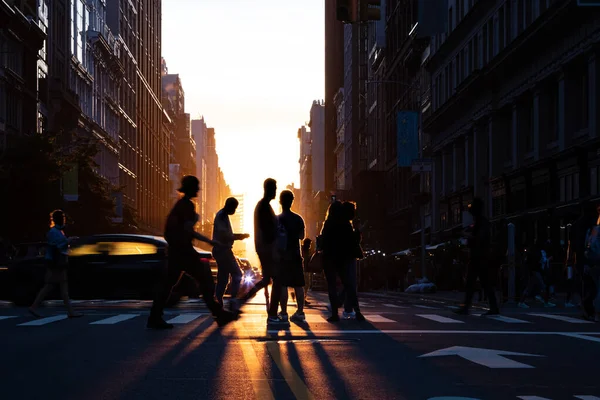 Silhouettes of people crossing a busy intersection on 5th Avenue in New York City with the light of sunset in the background