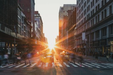People walking across a crosswalk at the busy intersection of 5th Avenue and 23rd Street in New York City with sunset light shining in the background
