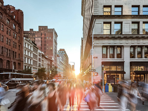 Crowds of people walking across a busy street intersection on 5th Avenue in New York City with sunlight shining in the background