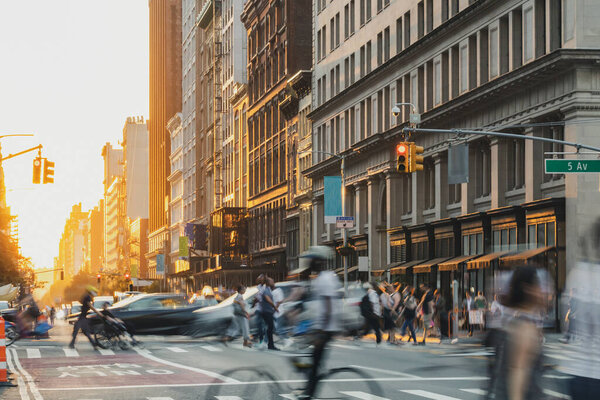 Crowds of people walking down the street at a busy intersection on 5th Avenue in New York City with sunset light shining in the background