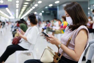 Hijyen maskesi takan, koluna yapıştırıcı yapıştırılmış, sandalyeye oturan, gözlem sırasında bulanık insanlar arasında akıllı telefon kullanan ya da Covid-19 aşısından sonra semptomları fark eden bir kadın..