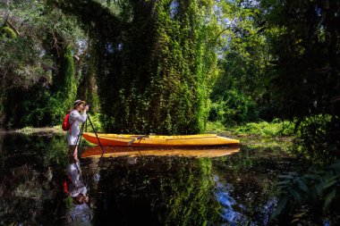 Woman researcher or botanist uses a camera on a tripod to photograph vegetation in a wetland forest. Female tourist stands in a swamp and takes pictures beside a canoe. Explorer working alone.