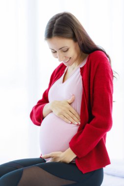 Pregnant woman sits on the bed in her bedroom, caressing her belly with a happy, smiling face. The joy of a new mother while touching her unborn baby and the arrival of a new born child.