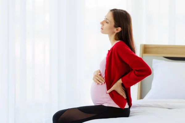 Pregnant woman sits on bed in bedroom, holding her back, showing pain or painful back. New moms with physical hurt, natural effects of pregnancy.