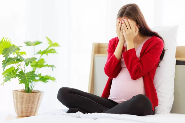 Pregnant woman sat on the bed in bedroom, covering her face with both hands, expressing a feeling of pressure and tensio.