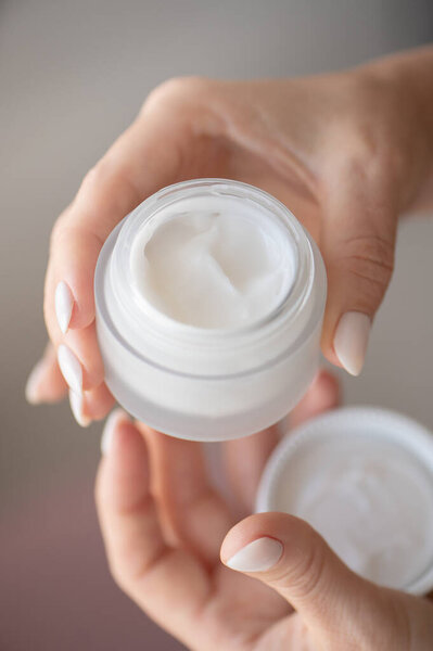 A woman's hands hold a white jar and use moisturizer. Taking care of her nails and clean, soft, smooth skin on her body. Front view. Close-up.