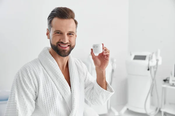 Attractive man in bathrobe shows cream jar in cosmetologycal salon