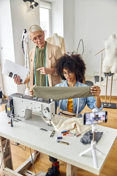 Middle aged lady with African-American dressmaker look at sleeve at sewing machine in studio