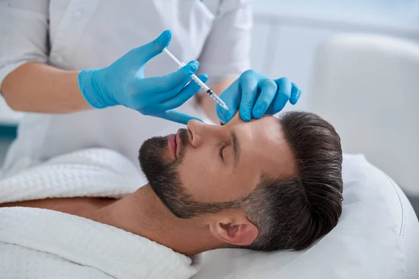 Beautician in blue gloves does injection at eyebrow lifting procedure to mature patient in clinic