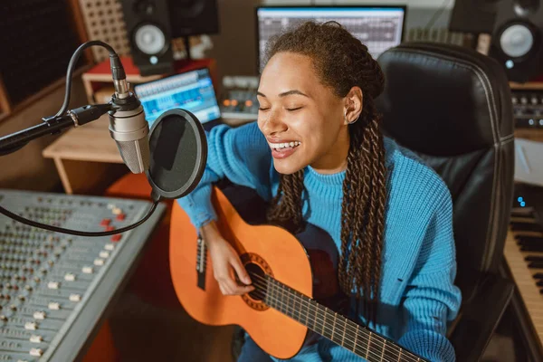 Enthusiastic multi-ethnic woman singer plays guitar and recordis song, sings into microphone in music recording studio