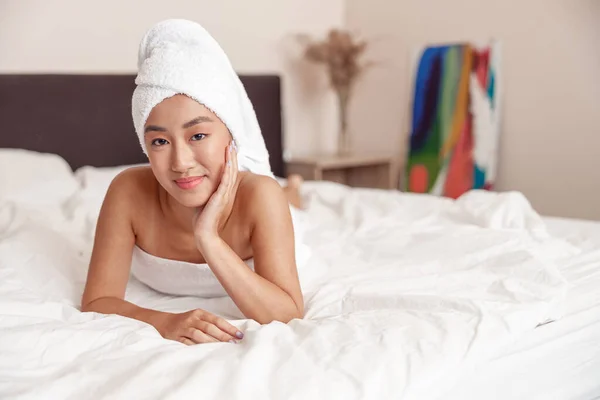 Female person with towel on head looking at camera and smiling while lying on bed at home