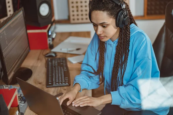 Concentrated multiethnic young woman, radio presenter, sound engineer wearing headphones typing text on laptop while working in broadcasting studio and sound recording stage