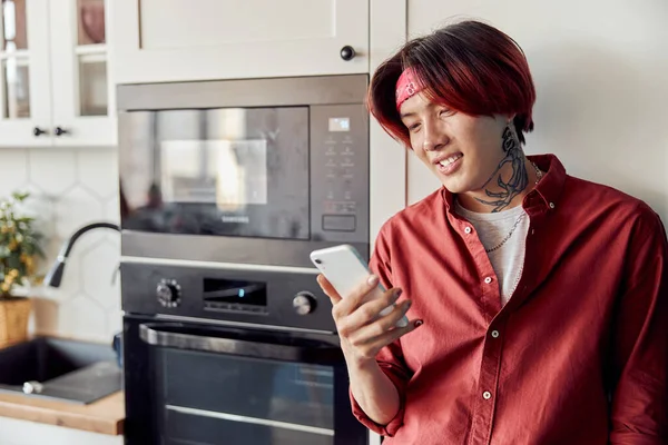 Waist up of smiling Asian guy typing on mobile phone in the kitchen near the microwave. Copy space