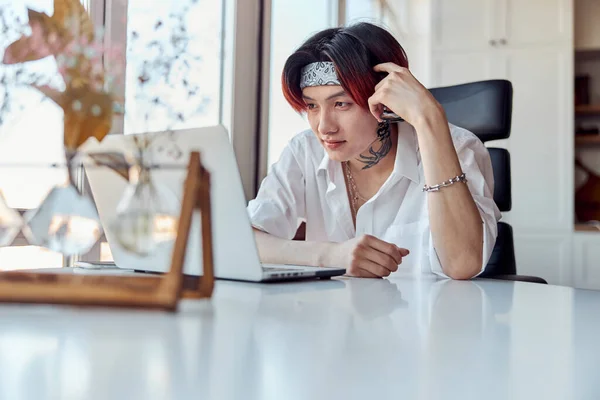 Stylish Asian guy looking at laptop screen at home with window on the background