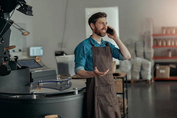 Handsome business owner talking phone on the background of roasting coffee machine on small factory