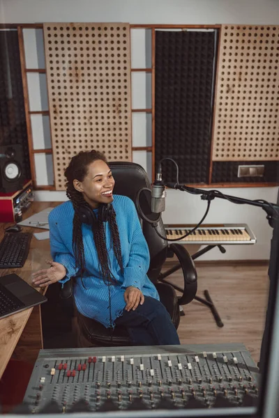 Multiethnic cheerful woman radio presenter with stylish dreadlocks sits in front of a mixing console with software and smiles, talking into microphone while broadcasting interesting program