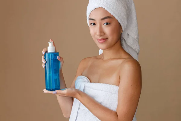 Female person with towel on her head looking at camera and smiling while holding cosmetic product. Isolated on light brown background