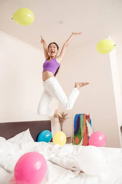 Happy excited lady jumping in the air and screaming with joy while having fun in bed with birthday balloons