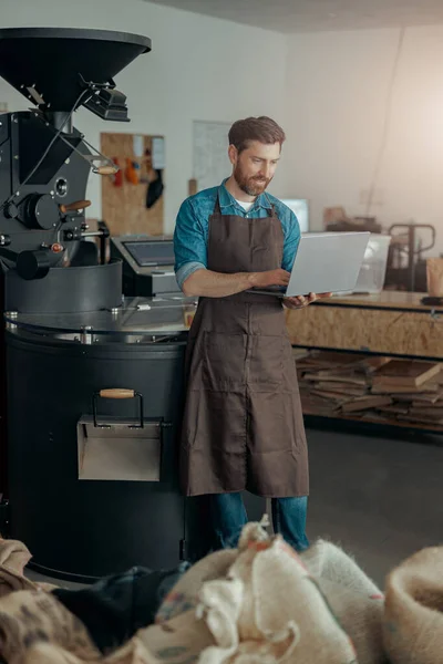 Handsome male worker on small coffee factory working laptop on background of coffee roasting machine