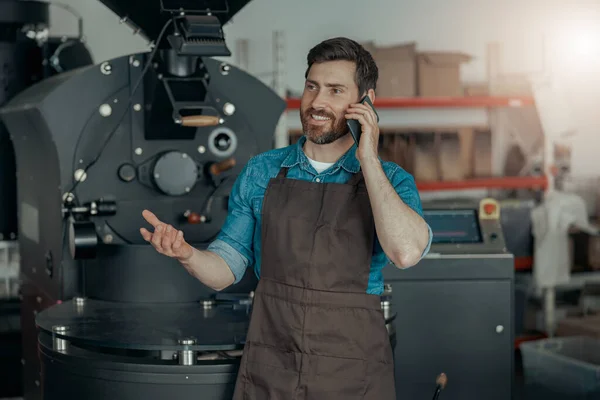 Business owner of small coffee factory talking phone on background of coffee roasting machine