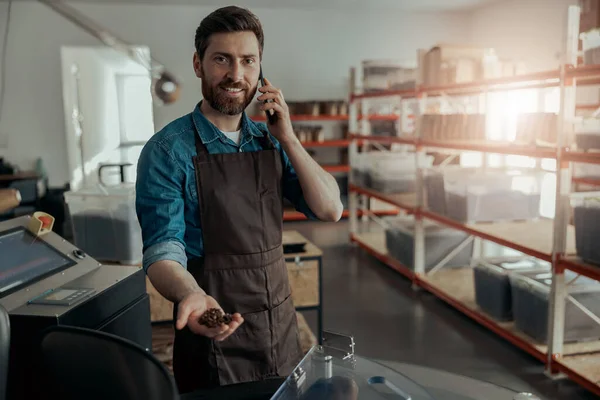 Barista on coffee factory talking phone on background of roasting machine and looking camera