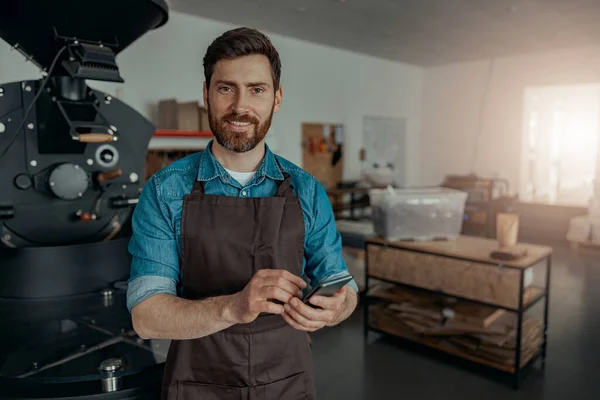 Business owner of small coffee factory using phone on background of coffee roasting machine