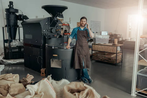Business owner of small coffee factory talking phone on background of coffee roasting machine
