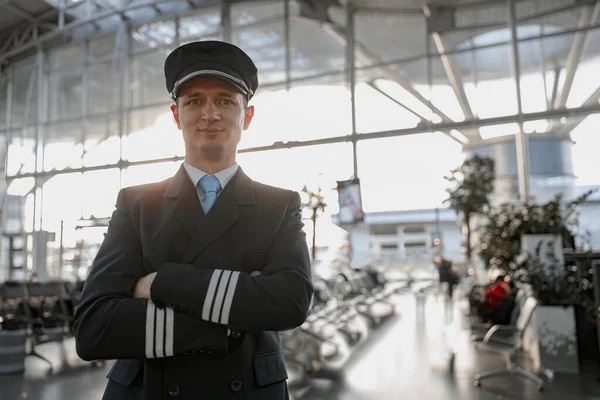 Waist up of smiling adult pilot in hat posing at camera in terminal with people on the background, copy space