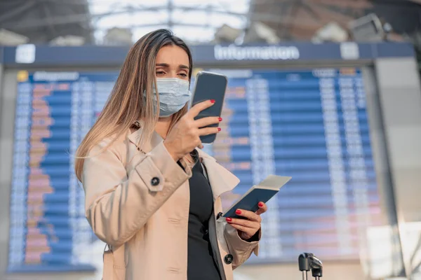 Pretty woman in protective mask waiting to board while looking at phone screen in airport