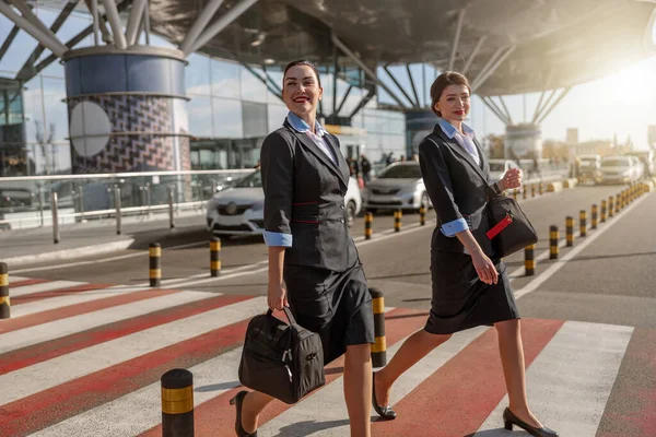 Side view of two young stewardesses with bags crossing the road near terminal in the airport