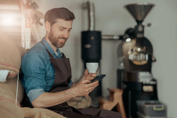 Positive worker drinking coffee sitting on bags with roasted coffee beans with phone during break