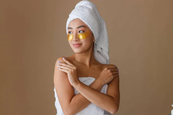 Female person with towel on her head using golden under-eye patches after shower. Isolated on light brown background