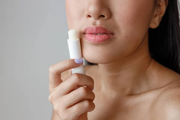 Close up of female person with plump lips holding moisturizing lip balm. Isolated on light gray background