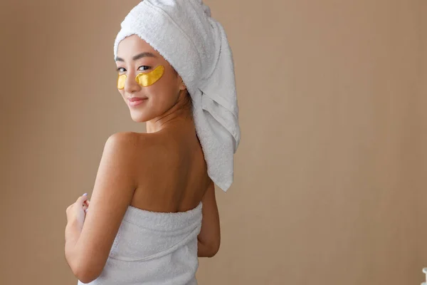 Asian female person with masks under lower eyelids wearing bath towel after shower. Isolated on light brown background