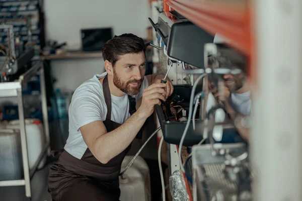 Smiling young man in uniform repairing coffee machine using screwdriver in a workshop