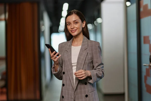 Portrait of attractive business woman with phone standing in modern coworking and looking camera