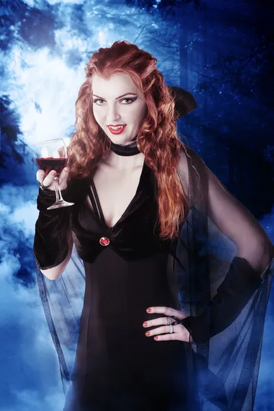Sexy vampire girl with glass of blood in the woods at night
