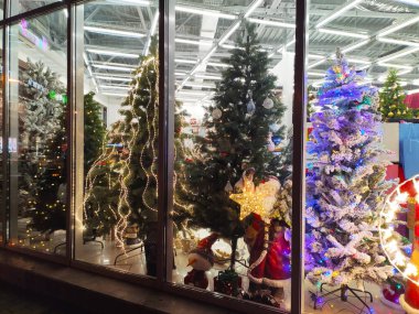 Vitrininde çeşitli Noel süsleri olan karlı yapay Noel ağaçları ve şeffaf cam arkasında soyut bulanık ışıklarla Noel Baba oyuncağı. Yeni Yıl Öznitelikleri