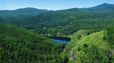 Lake Saryachik on the Ulagansky pass in Altai, view from above, a lake in a mountain lowland covered with dense forest, rocky slopes, mountain ranges in the distance, clear sky, summer, sunny