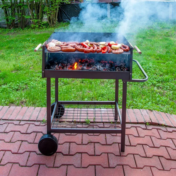 Assorted types of grilled meat with vegetables on the barbecue mobile grill with sausage, weisswurst, red pepper, bacon, onion and garlic with smoke and flames in the garden with green grass