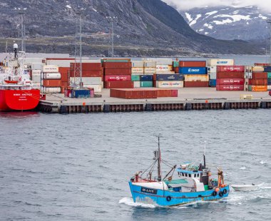 Small fishing returning to port in front of containers stacked up at the port in Nuuk with snow capped mountains in the background in Nuuk, Greenland on 20 July 2022